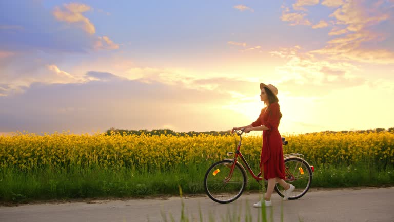 Tracking shot of a brunette in a red dress wearing a hat, as she walks her bike surrounded by snapdragon flowers, sunset in the background, slow motion