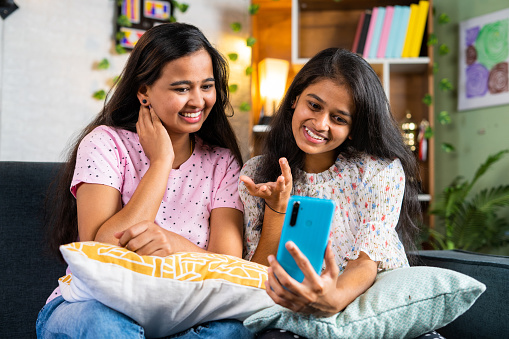 Happy smiling sibling girl sisters on sofa making video call on mobile phone at home - concept of relationship, wireless communication and cyberspace