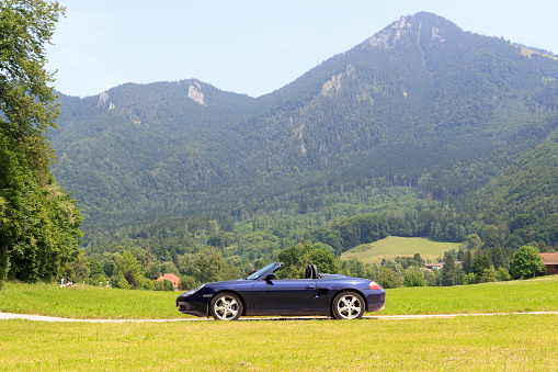 Ruhpolding, Germany - July 24, 2021: Blue roadster Porsche Boxster 986 with Bavarian Alps panorama at German Alpine Road. The car is a mid-engine two-seater sports car manufactured by Porsche.