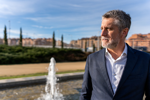 Thoughtful senior businessman with gray hair wearing formal suit standing near fountain and looking away while admiring amazing scenery