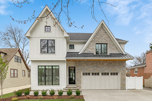 Oak Park, IL, USA - April 5, 2020: A new, white modern farmhouse with a dark shingled roof and black windows. The right side of the house has a light rock siding.
