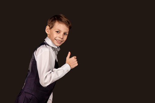 Schoolboy in school uniform showing thumb-up looking at camera on black isolated background. Good choice, well done. Stylish little boy in suit with bow tie, student posing in studio. Copy text space