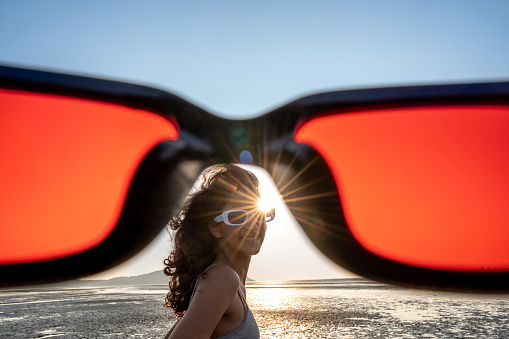 A young teenager wearing sunglasses and smiling on the beach seen from the point of view opf the photographer looking through red sunglasses.