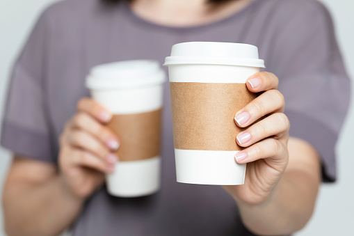 Unrecognizable caucasian female is  holding two disposable cups. One is for herself in the other cup is for a person in front of her.  She shows one of the cups to camera.