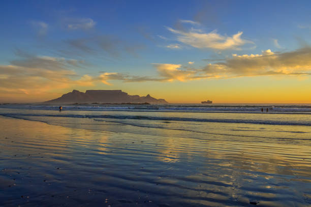 Authentic colors of dusk in the sky over the famous landmark of Table Mountain, Cape Town, as seen from the beach at Bloubergstrand to the north of the city. The waves coming in from the sea have been softened by a long exposure.