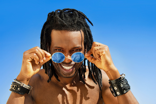 Cheerful man man with dreadlocks and blue sunglasses smiling under the sun at the beach, blue sky, sunny day