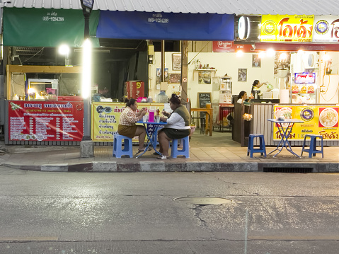Two thai women are sitting at table in front of restaurant and are eating. Table and plastic stools are placed on sidewalk.  Inside women are working in right background. Scene is on Wanghin Road in Bangkok Ladparo at early evening