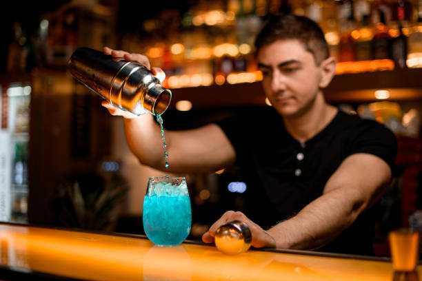 istock selective focus on glass with iced blue cocktail on bar and bartender pouring liquid into it from shaker 1458265622
