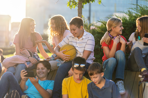 Group of happy teenage boys and girl sitting on the bench in city street while talking and using mobile phones