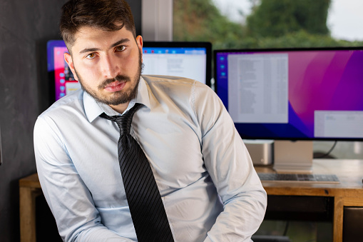 portrait of a serious young businessman, manager or office worker sitting with his arms crossed in front of his workstation