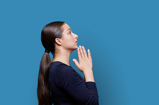 Young attractive female holding hands in prayer on blue studio background, profile view. Body language, please, sorry, help, plea, prayer, emotions, youth concept