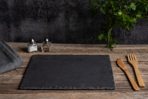 Empty black stone plate with wooden cutlery on a rustic old wooden table, next to salt, pepper and parsley leaves.