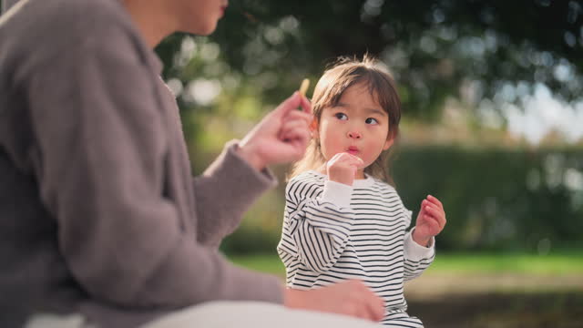 Small girl eating snack together with her mother in public park