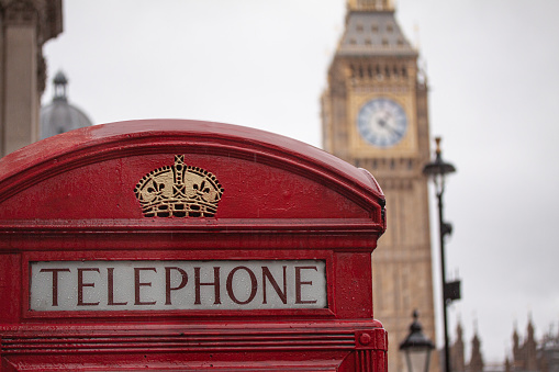 Traditional red telephone booth in London, England with blurred Big Ben clock by The Houses of Parliament  in the background.