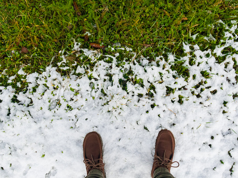 Man in brown boots standing on melting snow on green grass. Conceptual photograph of the movement toward spring.  Conceptual image about improving life. View from above. 
This image was taken with a mobile phone.