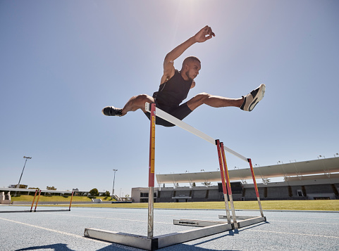 Male fit caucasian runner in sportswear running fast and jumping through hurdle at stadium track wide shot