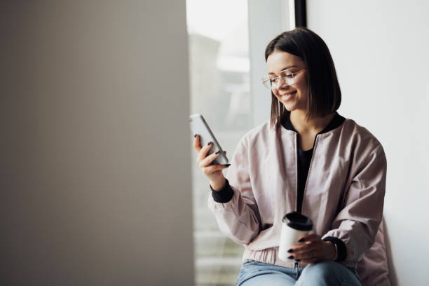 istock Young Woman in Glasses Drinking Coffee and Using Smartphone While Sitting on the Windowsill 1458299911