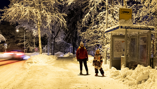 Finland in the city of Lahti, road safety. Young woman, Caucasian, with a child girl 7 years old on the street in winter. The family is waiting for the bus at the bus stop at night.