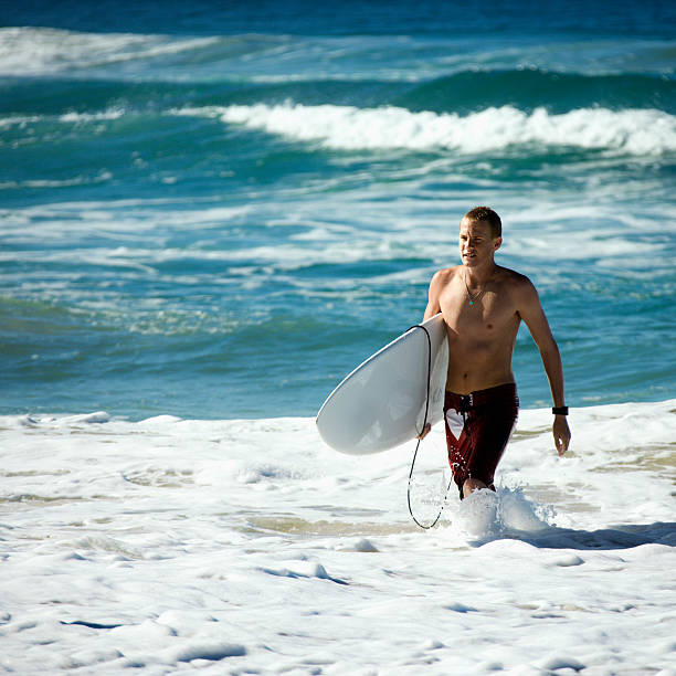 Atlantic Ocean at Taghazout, a tourist area in Morocco that is receiving considerable investment in new hotels, apartments etc. In the foreground is a surfer with a surf board walking into the surf across the wet sand.