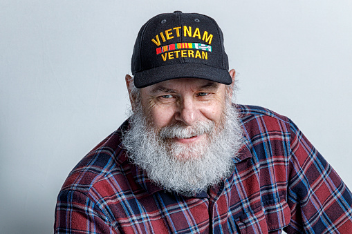 Portrait of a real life, real person senior adult man United States Navy USA Vietnam War military veteran with a full, bushy, gray beard and a quiet smile. He's wearing a commemorative Vietnam War Veteran's baseball cap style hat.