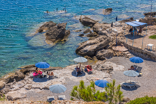 Greece. Rhodes. 08.25.2022. Beautiful view of rocky coast against backdrop of turquoise water in Mediterranean Sea and sun loungers with people on rock beach. .