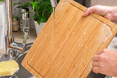 A man washes wooden bamboo cutting board in the kitchen sink under running water. Gentle hand washing of a wooden cutting board. Cleaning dirty kitchen wood products.