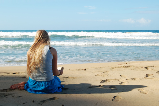 A young woman in a blue skirt and bare feet sits on a sandy beach and reads.