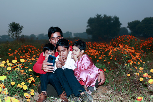 Indian family sitting together in flower field during springtime peacefully and watching something on mobile phone portrait.