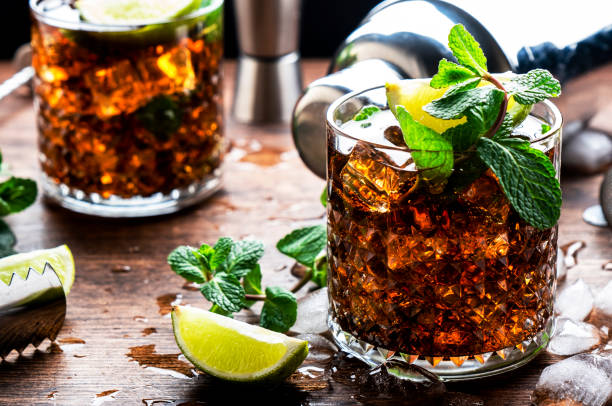 Rum cola cocktail with strong alcohol and ice, garnished with mint and lime in glass. Wooden background, hard light stock photo