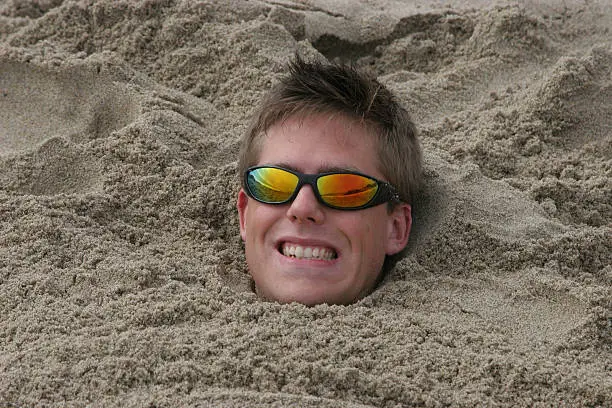 20-30 year old man up to his neck in the sand. Smiling and wearing sunglasses.