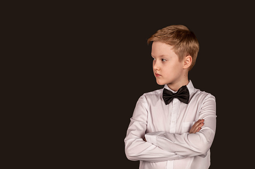 Portrait serious schoolboy in white shirt with bow tie on black isolated background. Fashionable little boy, student crossed hands looking away in studio. Education concept. Copy text space for ad
