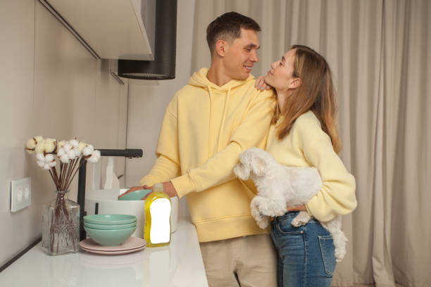 Happy couple in yellow hoodies, man washing the dishes, woman holding little white dog Happy couple in yellow hoodies, man washing the dishes, woman holding little white dog laundry husband housework men stock pictures, royalty-free photos & images