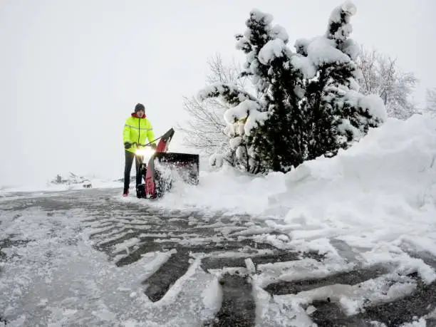 Young man wearing a high visibility jacket removing and clearing snow of the road, driveway and footpath, using a red snowblower after the snow storm.
