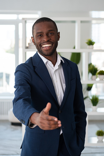 Smiling African American businessman wearing formal suit is standing stretching out hand for handshake at office workplace in background. Concept of model, business person, director, boss, greeting