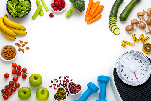 Top view of an assortment of healthy fruits and vegetables, a weight scale, a measure tape and dumbbells arranged side by side on a frame shape leaving a useful copy space at the center of the image on a white background