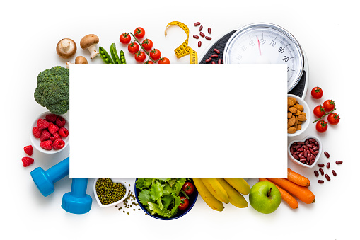 Top view of an assortment of healthy fruits and vegetables, a weight scale, a measure tape and dumbbells isolated on white background with a blank copy space at the center of the image