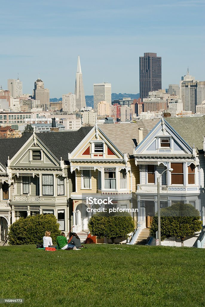 Girls Sitting in Front of the "Painted Ladies" Victorian Houses Three teenage girls sitting in Alamo Square Park in front of ""The Painted Ladies"" Victorian houses with the San Francisco skyline in the background. Architecture Stock Photo