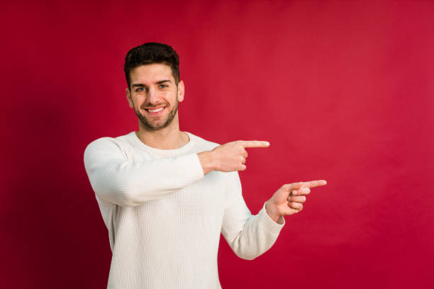 Cheerful young man pointing to the left with both hands on red background