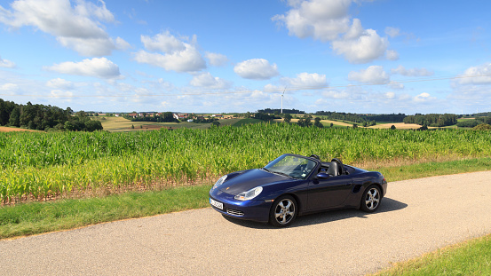 Schopfloch, Germany - July 31, 2021: Blue roadster Porsche Boxster 986 with corn field panorama at Romantic Road. The car is a mid-engine two-seater sports car manufactured by Porsche.