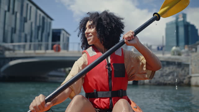 Black woman, kayak and water sports during travel outdoor for adventure and exercise. Happy fitness female on city lake with a boat for rowing or kayaking with urban buildings on holiday or vacation