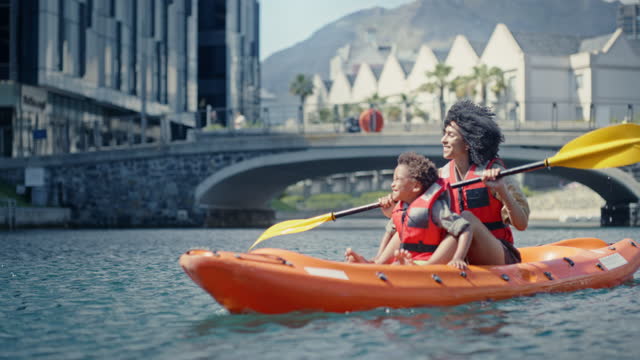 Kayak adventure, black family and pointing for travel outdoor during exercise and water sport. Woman or mother and kid on lake with a boat for rowing on bonding holiday or vacation in Cape town