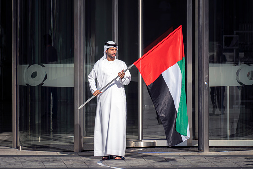 Dubai, UAE - November 28, 2022: Arab man with a traditional white shirt waves the flag of the UAE