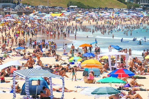Large crowd of people on Bondi Beach in the Eastern Suburbs of Sydney on 2 January 2023.  The red and yellow coloured uniforms and surfboards of surf lifesavers are visible in the crowd, as is their yellow and red tent. The red and yellow flags indicate the area where it is safe to swim.  This image was taken at the northern end of the beach in the early afternoon.