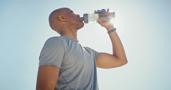 Drinking water, fitness and black man after workout, running training and exercise outdoor in sun. Blue sky, sport runner and water drink of a athlete with a bottle taking a sports break in nature