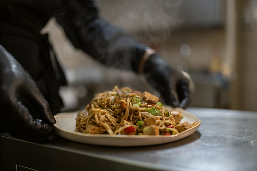 A chef from Malaysia in a black uniform cooks pasta and prepares pasta and vegetables in a wok.