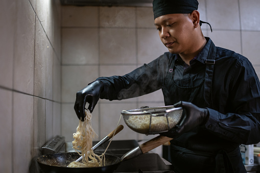 A chef from Malaysia in a black uniform cooks pasta and prepares pasta and vegetables in a wok.