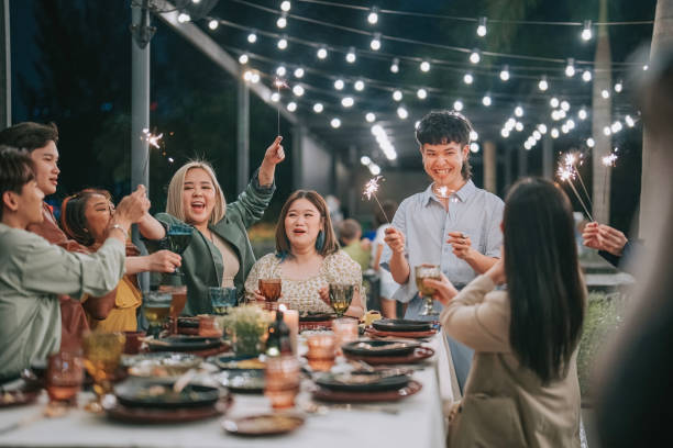 Asian Chinese Friends Outdoor Dining celebrating with sparkler night social gathering enjoying drink and food Asian Chinese Friends Outdoor Dining at night social gathering enjoying with sparkler drink and food celebrating with sparkler fireworks on new year's eve having fun together in patio party - Celebration and holidays gay dinner table stock pictures, royalty-free photos & images