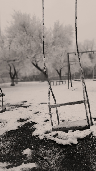 Black and White Vertical photo of Abandoned Broken Frozen Metal Swing on forgotten Snow Covered Playground in Winter