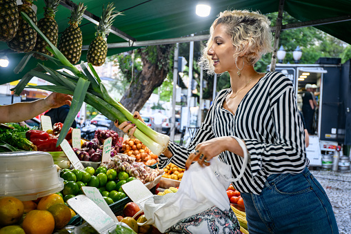 Beautiful young blonde woman smiling receiving leeks to put in the reusable bag at the market stall in a farmer's market in a plaza during a late summer afternoon