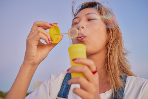 Asian, nature and summer bubbles with girl for vacation, youth and wellness on peaceful evening in Japan. Calm, relax and peace of young woman enjoying outdoor sunset leisure blowing soap bubbles.